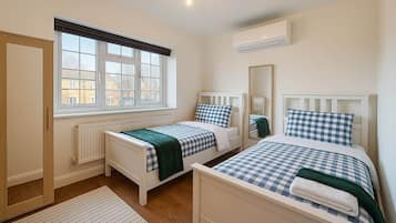 A bedroom with two single beds, a window with blinds, a radiator, and an air conditioning unit.