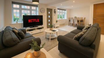 A modern living room with a flat-screen TV, two sofas, a coffee table, and a dining area in the background.
