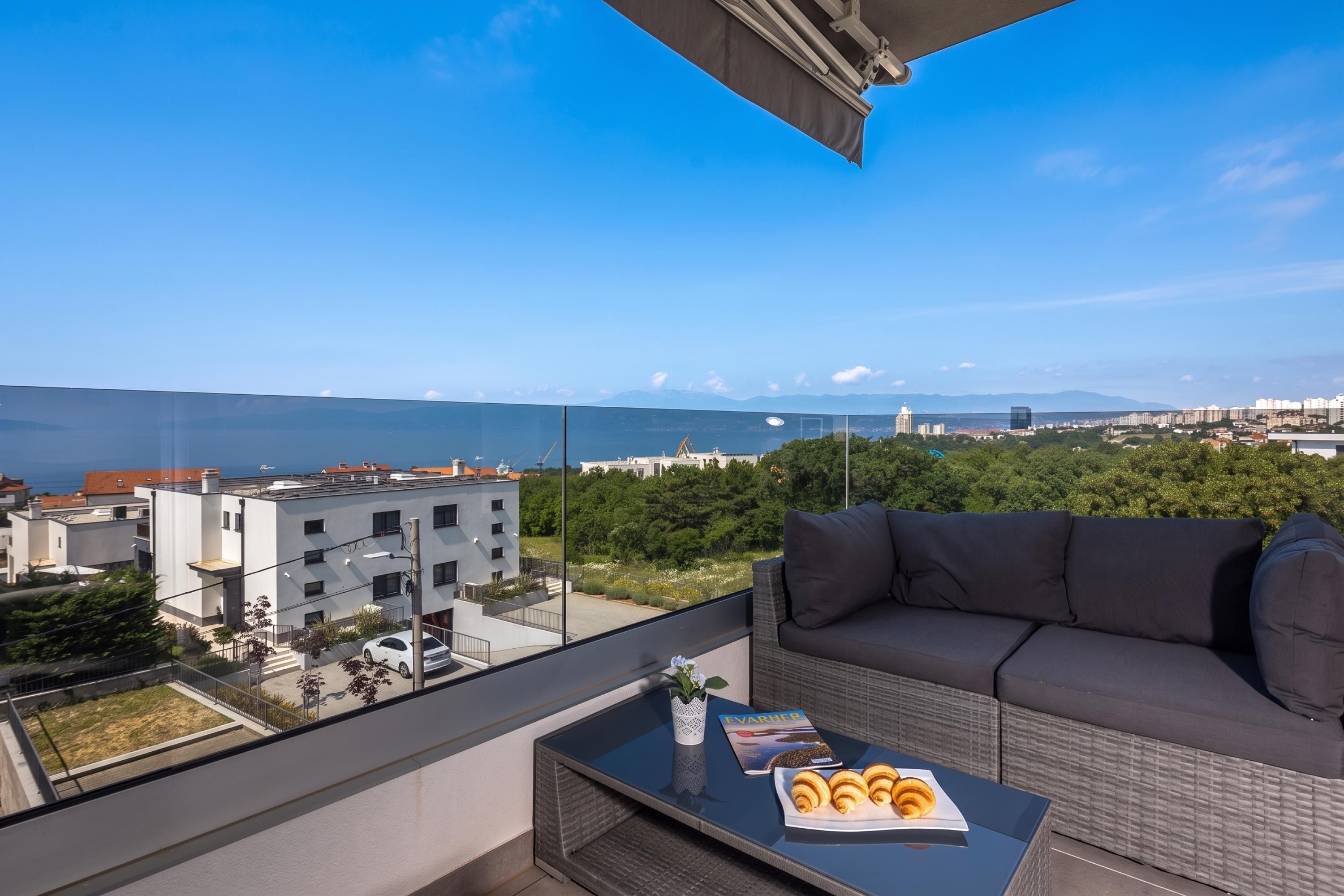 A balcony with a sofa and a table, overlooking a cityscape and the sea.