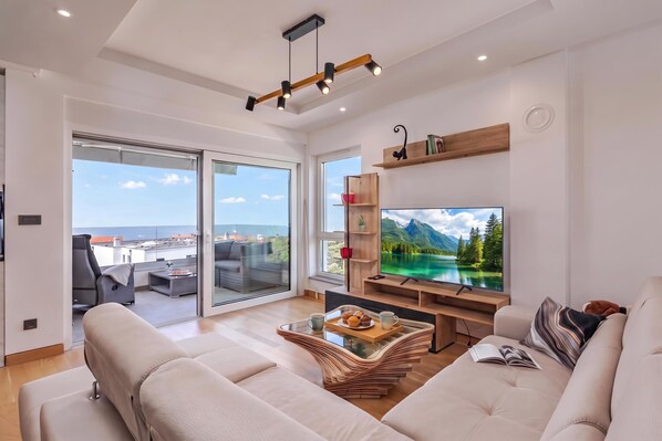 A modern living room with a large flat-screen TV, a sofa, a coffee table with a tray of muffins, and a view of a mountainous landscape through the glass doors. - Seaview Apartment Kostrena Two (Kostrena)