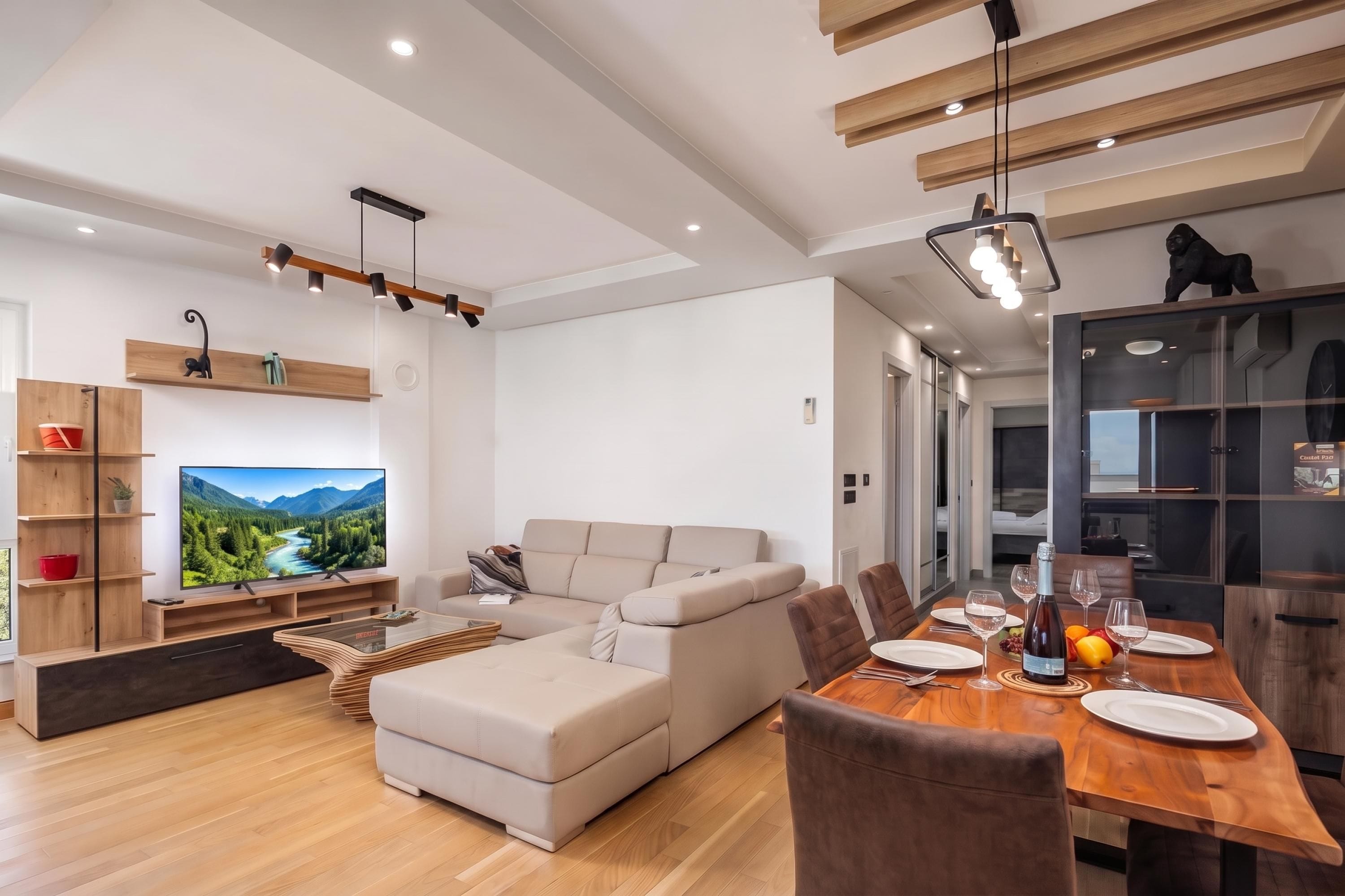 A modern living room with a large flat-screen TV, a sofa, a dining table set for four, and a kitchen area with cabinets and a refrigerator.