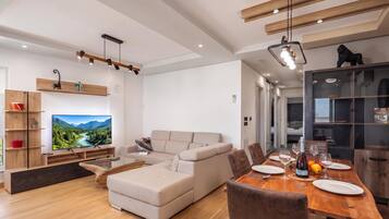 A modern living room with a large flat-screen TV, a sofa, a dining table set for four, and a kitchen area with cabinets and a refrigerator.