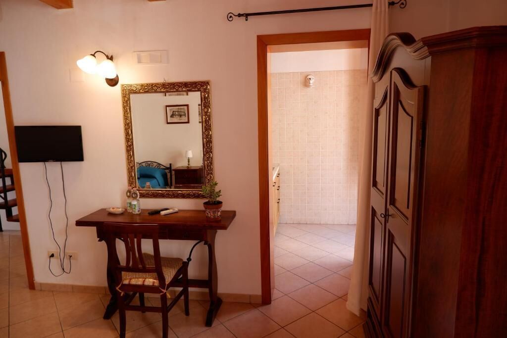 A room with a wooden desk, chair, and a large mirror with a decorative frame. There is a television mounted on the wall and a door leading to another room.