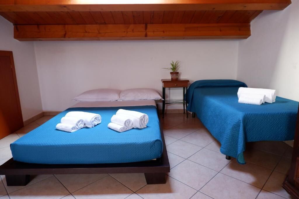 Ein Zimmer mit zwei Betten, jeweils mit blauer Bettdecke und weißen Handtüchern. Es gibt einen kleinen Tisch mit einer Pflanze und einer Schublade.