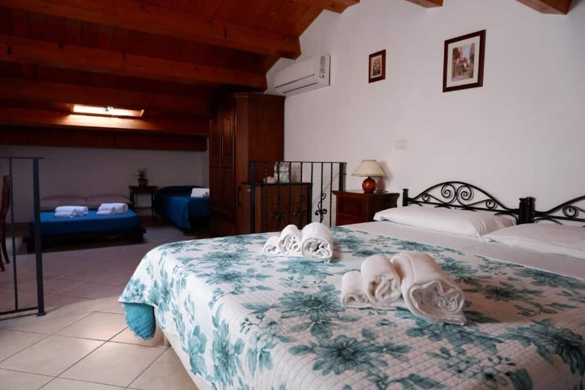 Ein Hotelzimmer mit einem großen Bett, einem kleineren Bett, einem Schreibtisch und einem Stuhl. Es gibt ein Fenster und eine Klimaanlage an der Wand.