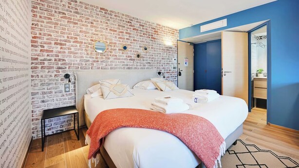 A bedroom with a brick-patterned wall, a large bed with white linens, a red throw blanket, and a small bedside table.
