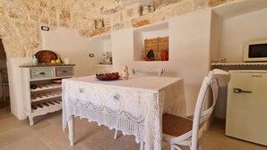 Uma área de refeições acolhedora, com uma mesa branca e toalha rendada, uma cadeira branca, um pequeno frigorífico e uma parede de pedra.