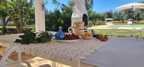 Mesa de comedor al aire libre con mantel de encaje blanco, una jarrilla, un bol de aperitivos y una vista a una piscina y árboles al fondo.