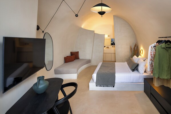 A modern bedroom with a curved ceiling, a bed, a seating area, a TV, and a wardrobe. - Calissa Suites by Ananda (Santorini)