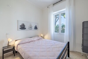 Un dormitorio con una cama, una mesita de noche con lámpara, una ventana con cortinas y una obra de arte colgada en la pared.