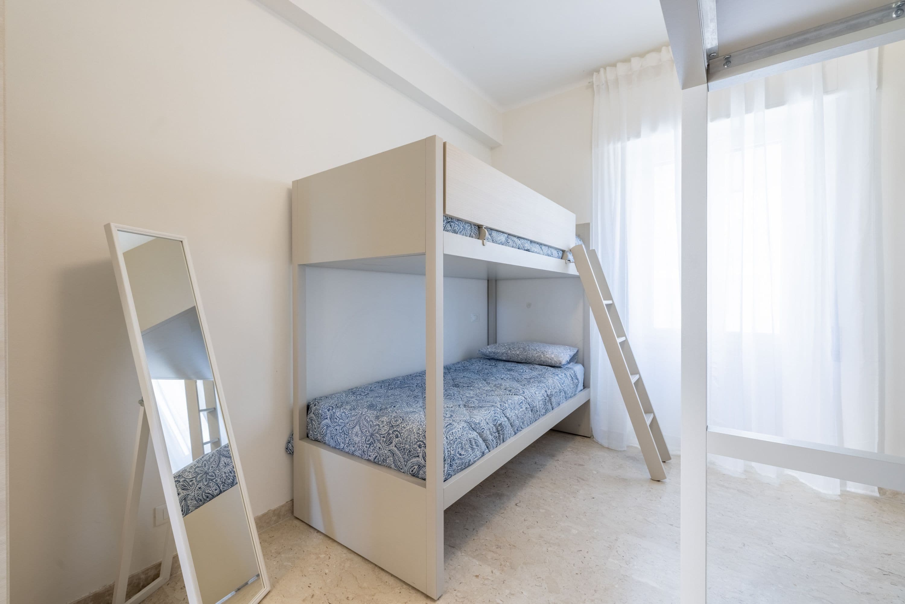 A bunk bed with a ladder, a mirror, and a window with sheer curtains.