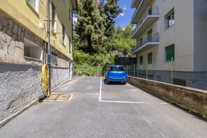Une rue étroite où une voiture bleue est garée sur le côté droit, bordée de bâtiments dotés de balcons et d’un mur de pierre sur le côté gauche.