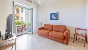 A living room with a sofa, a TV on a table, a painting on the wall, and a view of a balcony with outdoor furniture. - Next to the Beach by Wonderful Italy (Ospedaletti)