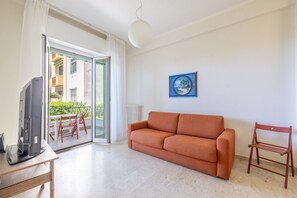 Een woonkamer met een bank, een televisie op een tafel, een schilderij aan de muur en uitzicht op een balkon met tuinmeubilair.