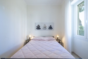 Una cama bien hecha con un colcha de tonos claros, una obra de arte en la pared con piedras apiladas, y una ventana con vista a árboles.