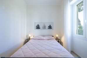 Una cama bien hecha con un colcha de tonos claros, una obra de arte en la pared con piedras apiladas, y una ventana con vista a árboles.