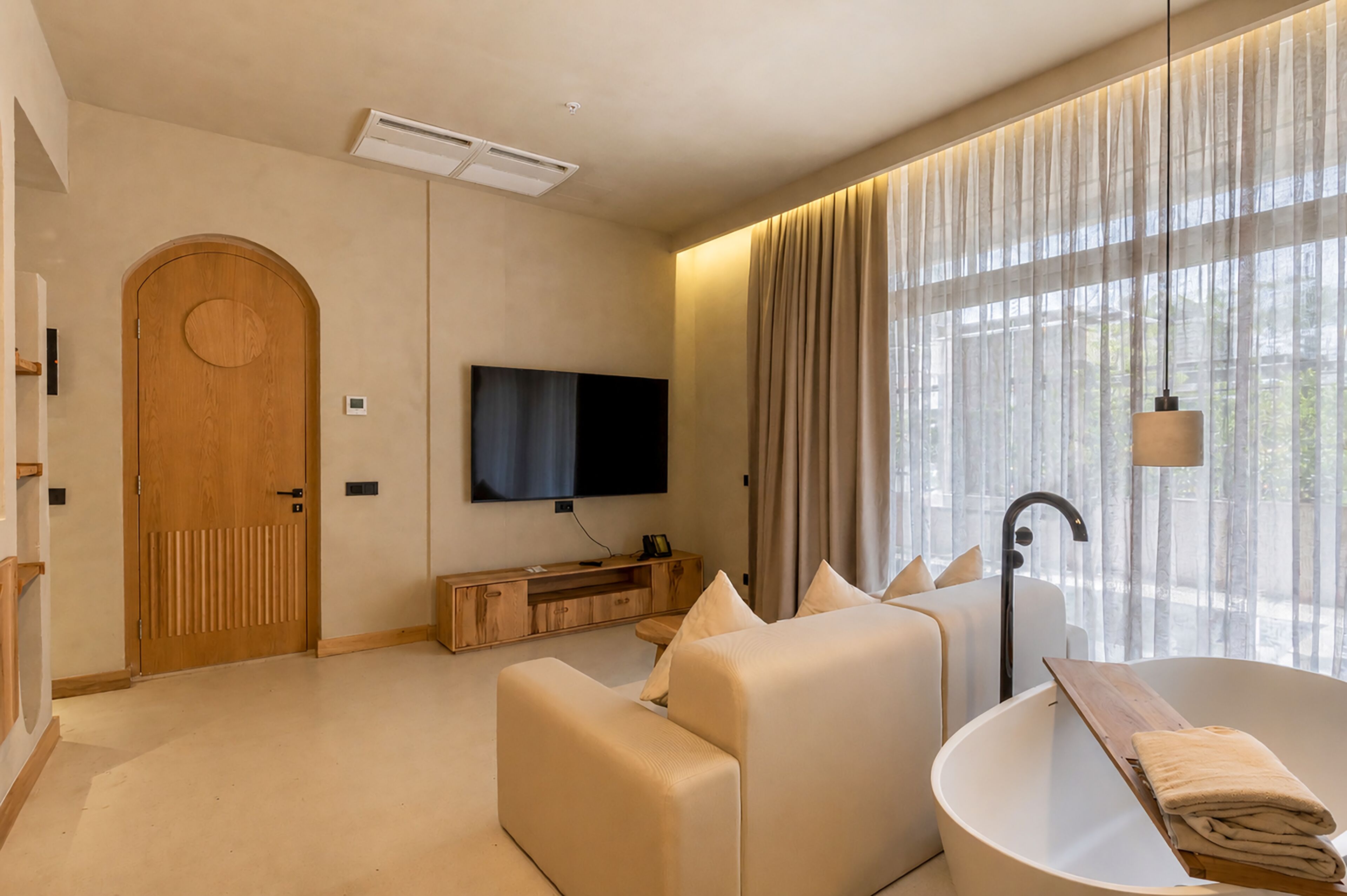 Une chambre d’hôtel moderne avec une porte en bois, un téléviseur à écran plat, un canapé, une baignoire et une grande fenêtre avec des rideaux.