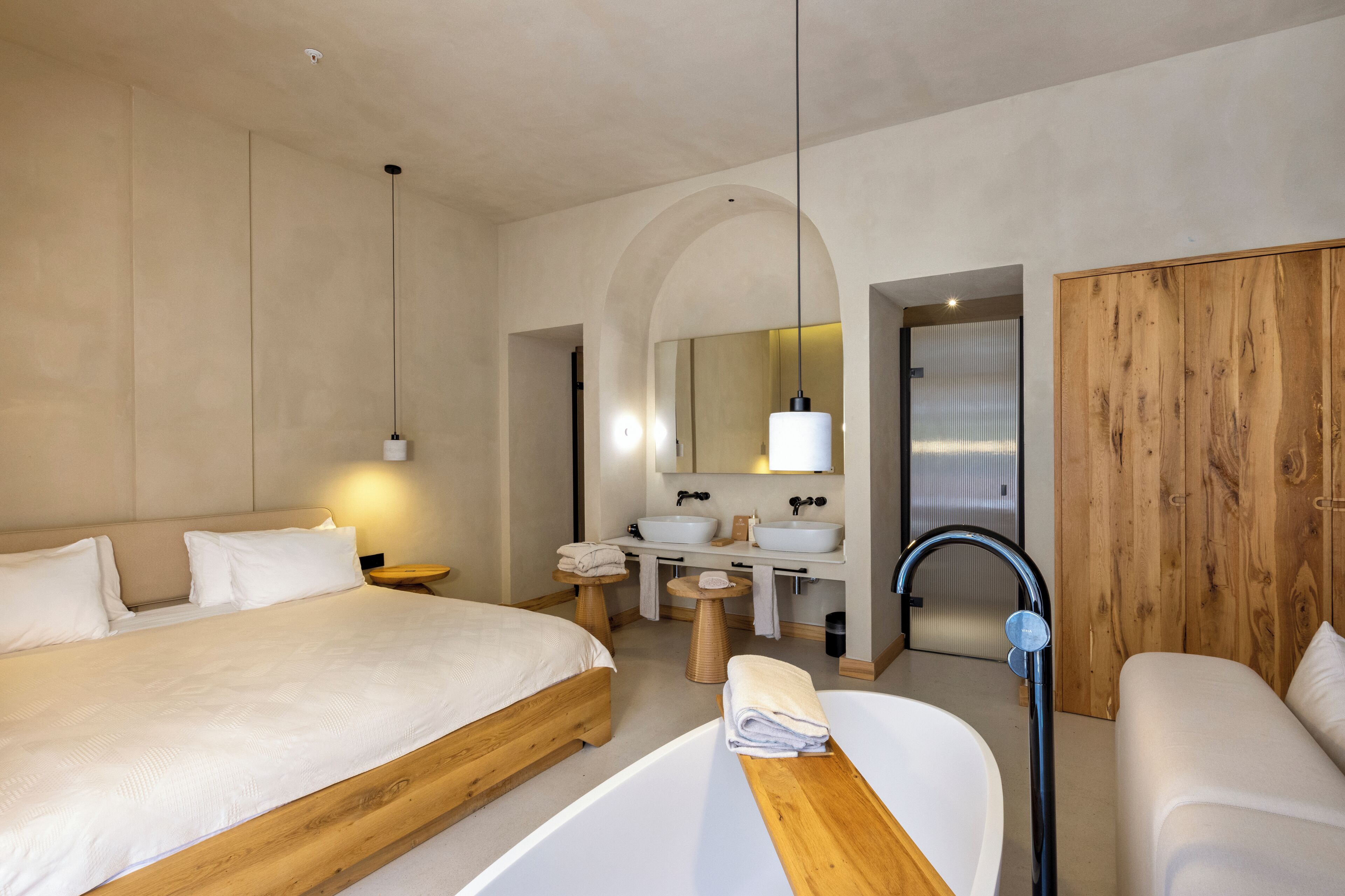 Habitación de hotel moderna con una cama grande, un sofá independiente, una bañera, un tocador con espejo y un armario de madera.