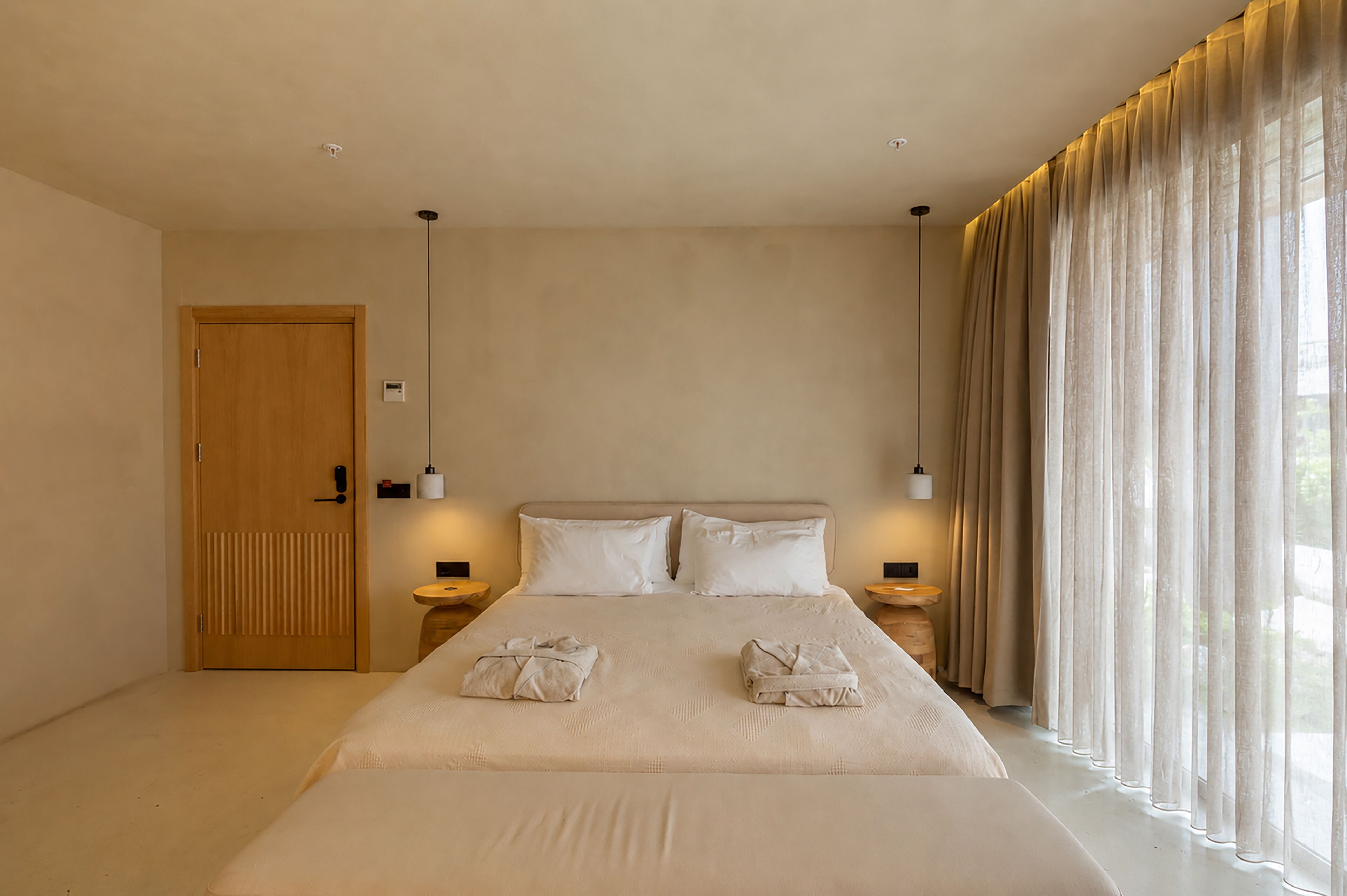 A hotel room with a large bed, two bedside tables, a wooden door, and sheer curtains.