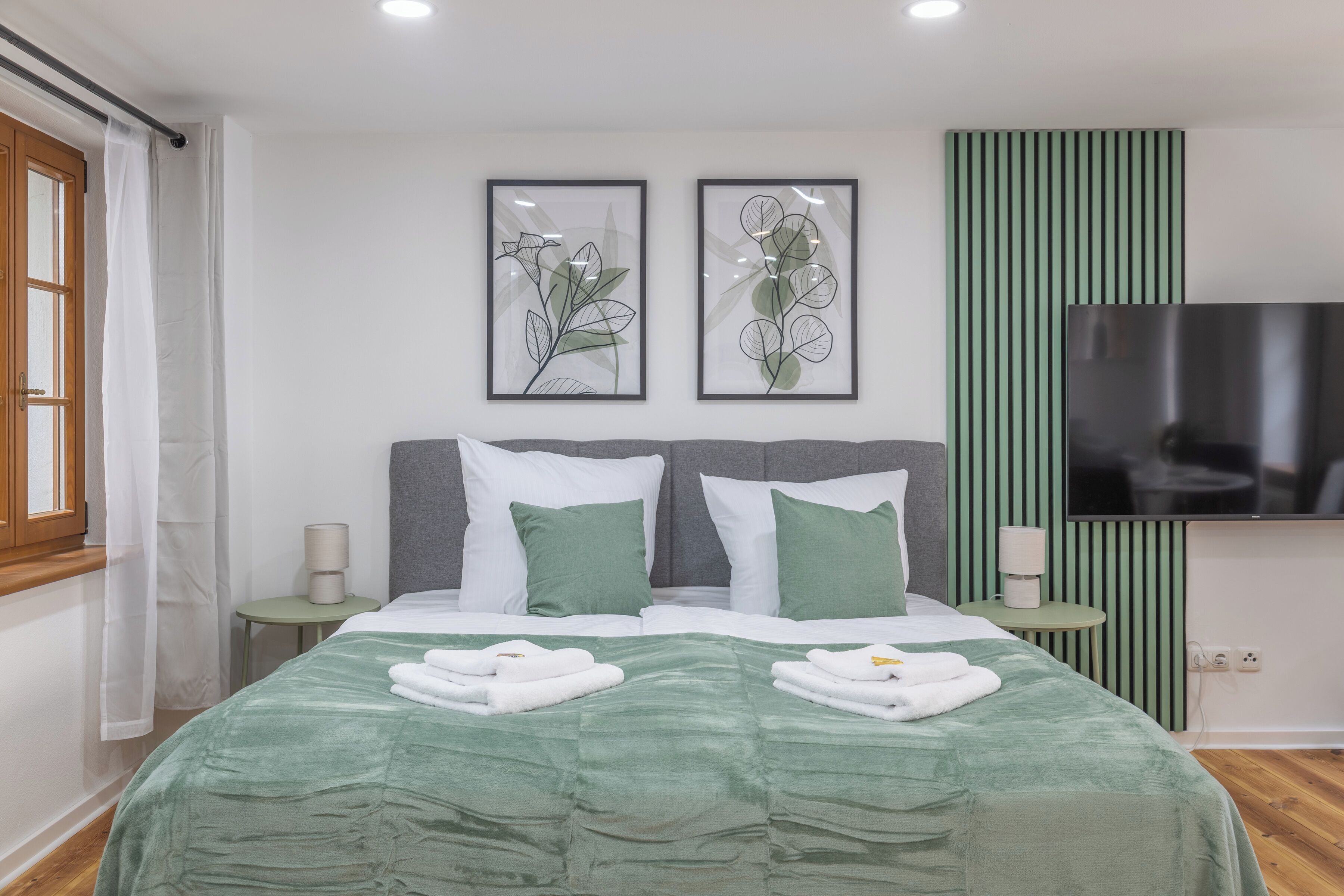 Una habitación de hotel moderna con una cama grande, ropa de cama verde, dos impresiones enmarcadas de motivos botánicos, un televisor de pantalla plana y una pared de acento verde.