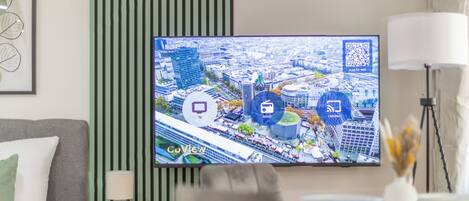 Quarto de hotel moderno com uma televisão grande exibindo uma vista urbana, uma mesa redonda pequena com abajur, parede com listras verdes e uma obra de arte emoldurada.