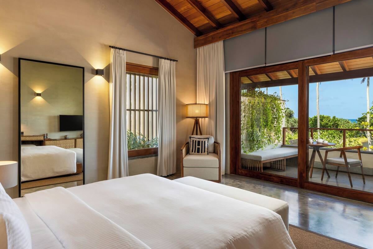 A hotel room with a large bed, a desk, a chair, a lamp, and a view of the outdoors.