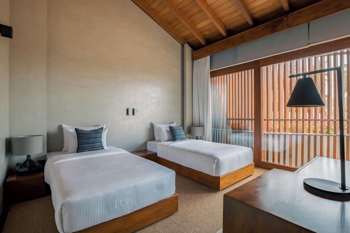 A hotel room with two beds, wooden headboards, a large window with blinds, a ceiling fan, and a nightstand with a lamp.