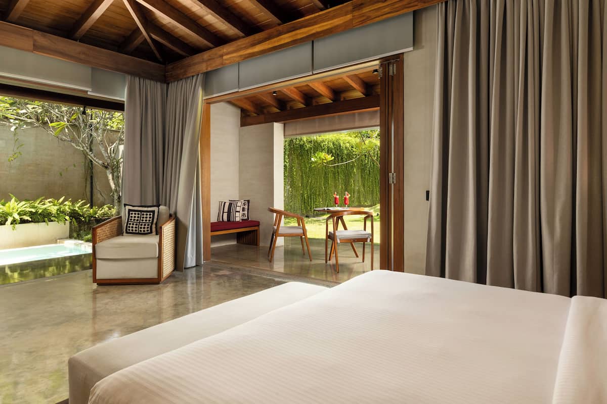 A modern bedroom with a large bed, a wooden ceiling, a seating area with a table and chairs, and a view of a swimming pool and greenery.