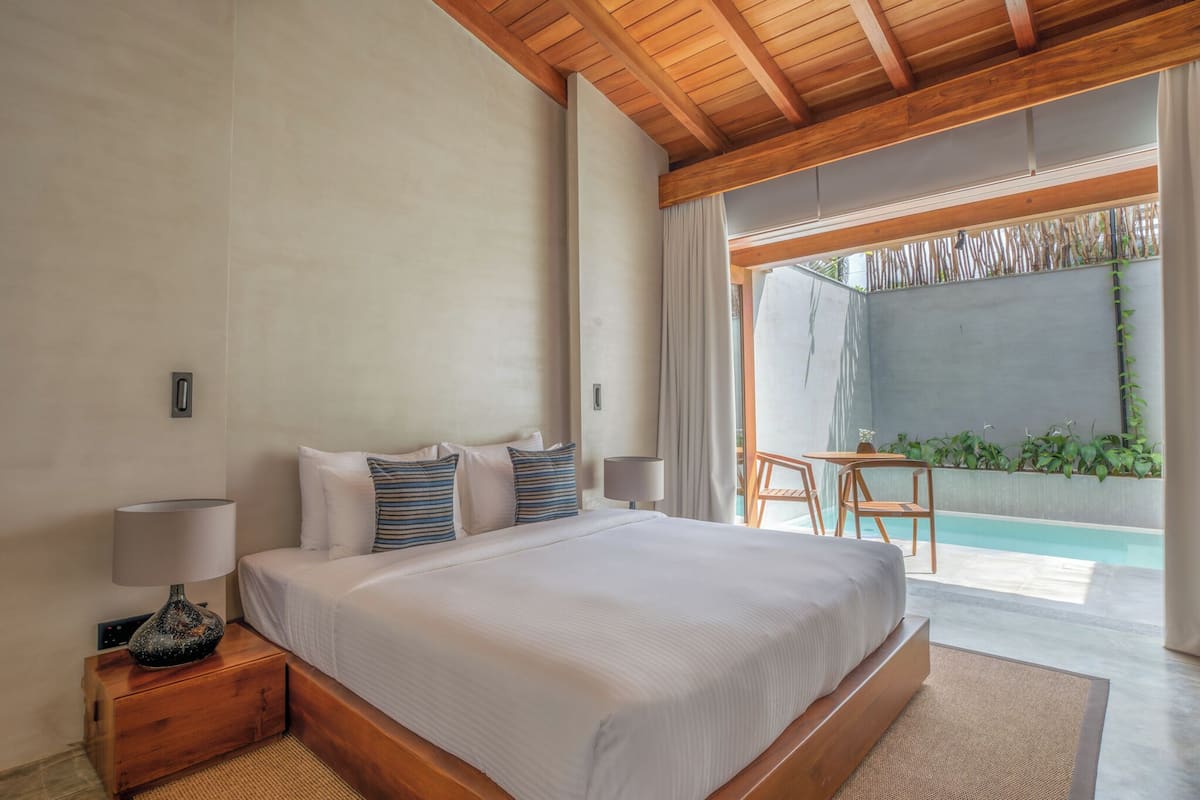 A bedroom with a large bed, bedside lamps, a wooden nightstand, and a view of an outdoor pool area.
