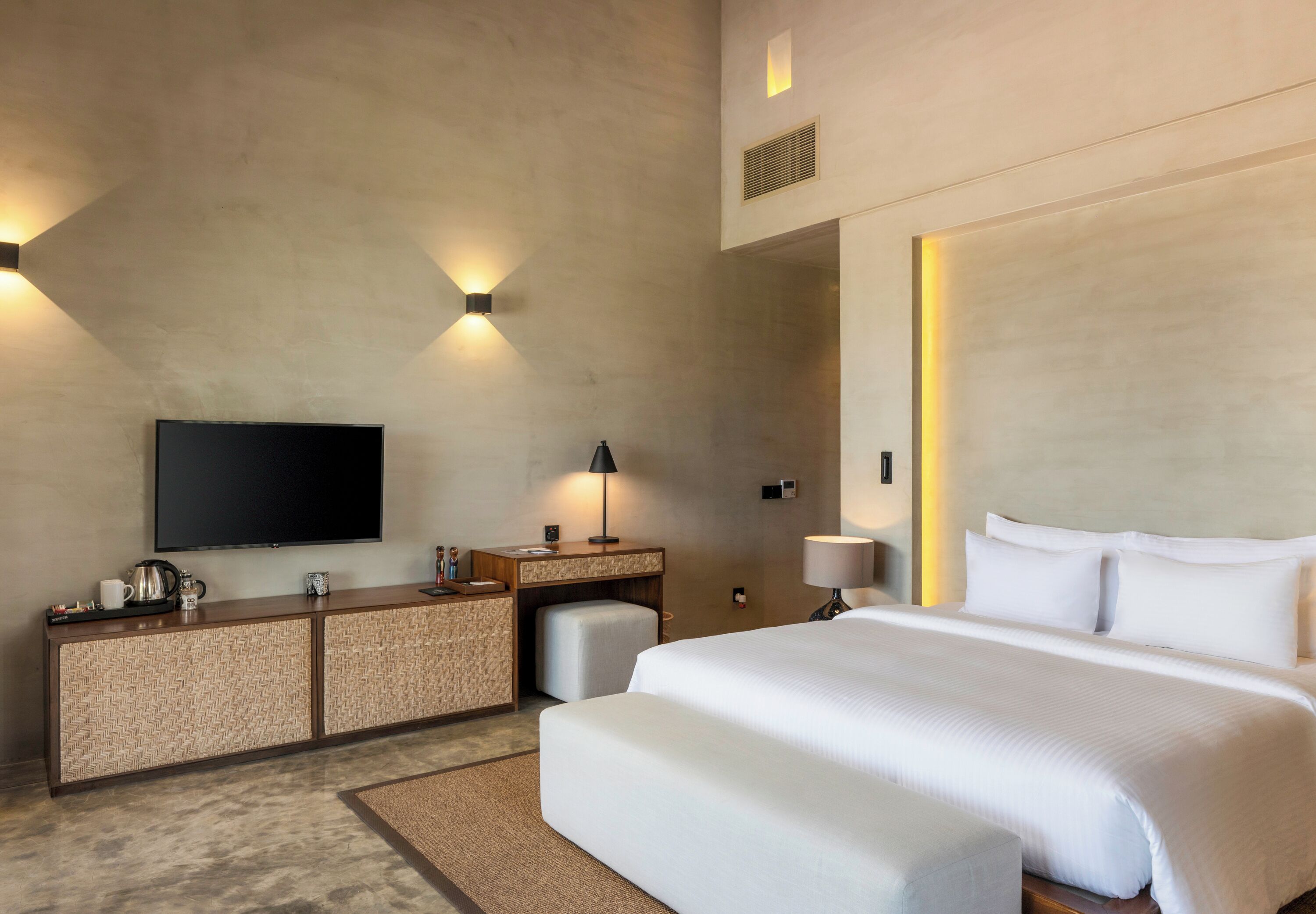 A modern hotel room with a large bed, a flat-screen TV mounted on the wall, a desk with a lamp, and a small seating area.