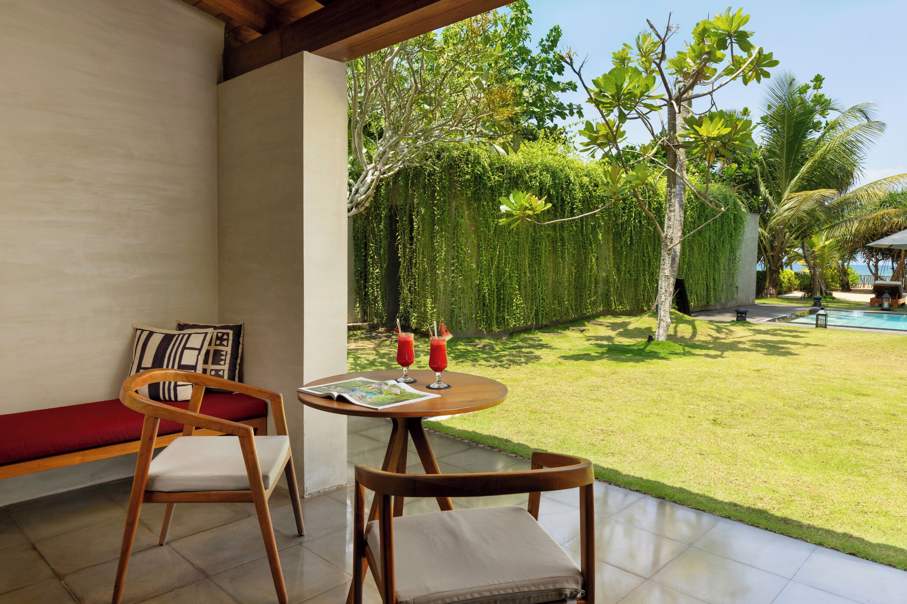 A patio with a wooden table and chairs, a red sofa, and a view of a lush garden with a pool.