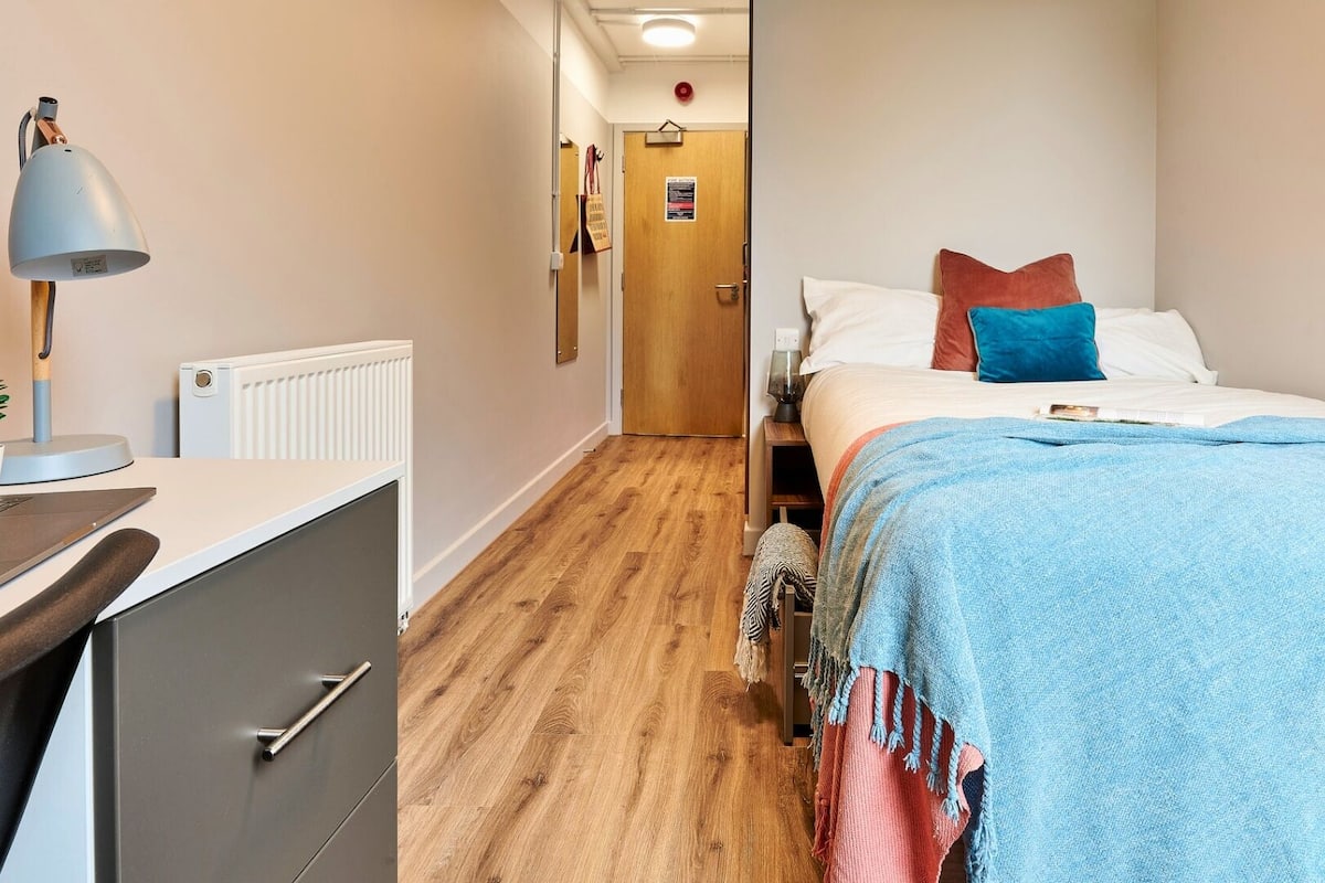 A bedroom with a bed, a desk with a lamp, a radiator, and a wooden floor.