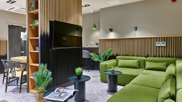 A modern lounge area with a green sofa, a wooden bookshelf, a flat-screen TV, and a small table with a magazine.