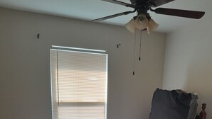 Un dormitorio con una cama individual, un ventilador de techo, una ventana con persianas y un caja en el suelo.