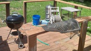 Un patio trasero con una terraza de madera, una parrilla, una funda para la parrilla, un balde azul, un tanque de propano y una caseta pequeña para perro.