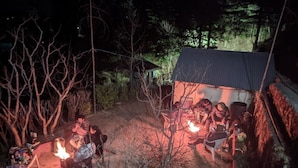 Ryhmä ihmisiä oli kokoontunut nuotion ympärille metsäisellä alueella, ja taustalla näkyi pieni rakennus.