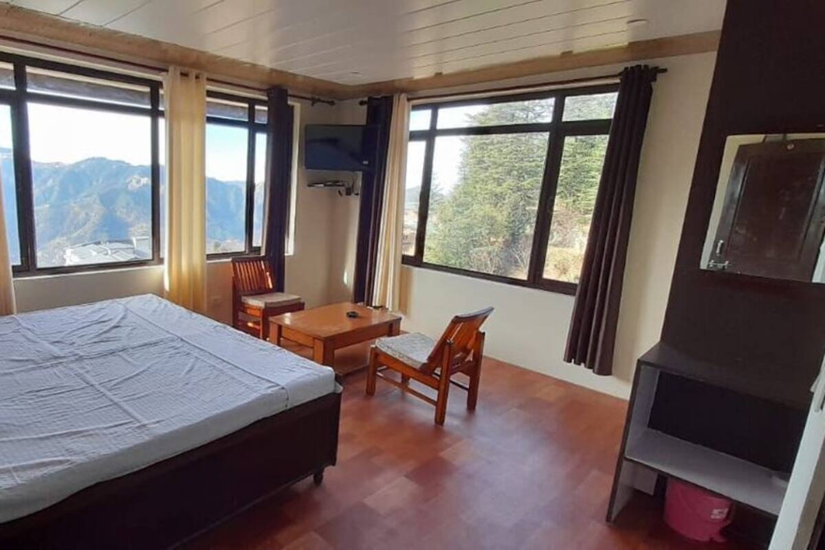 Quarto com cama, escrivaninha e cadeira. Há janelas amplas com cortinas e uma TV fixada na parede.