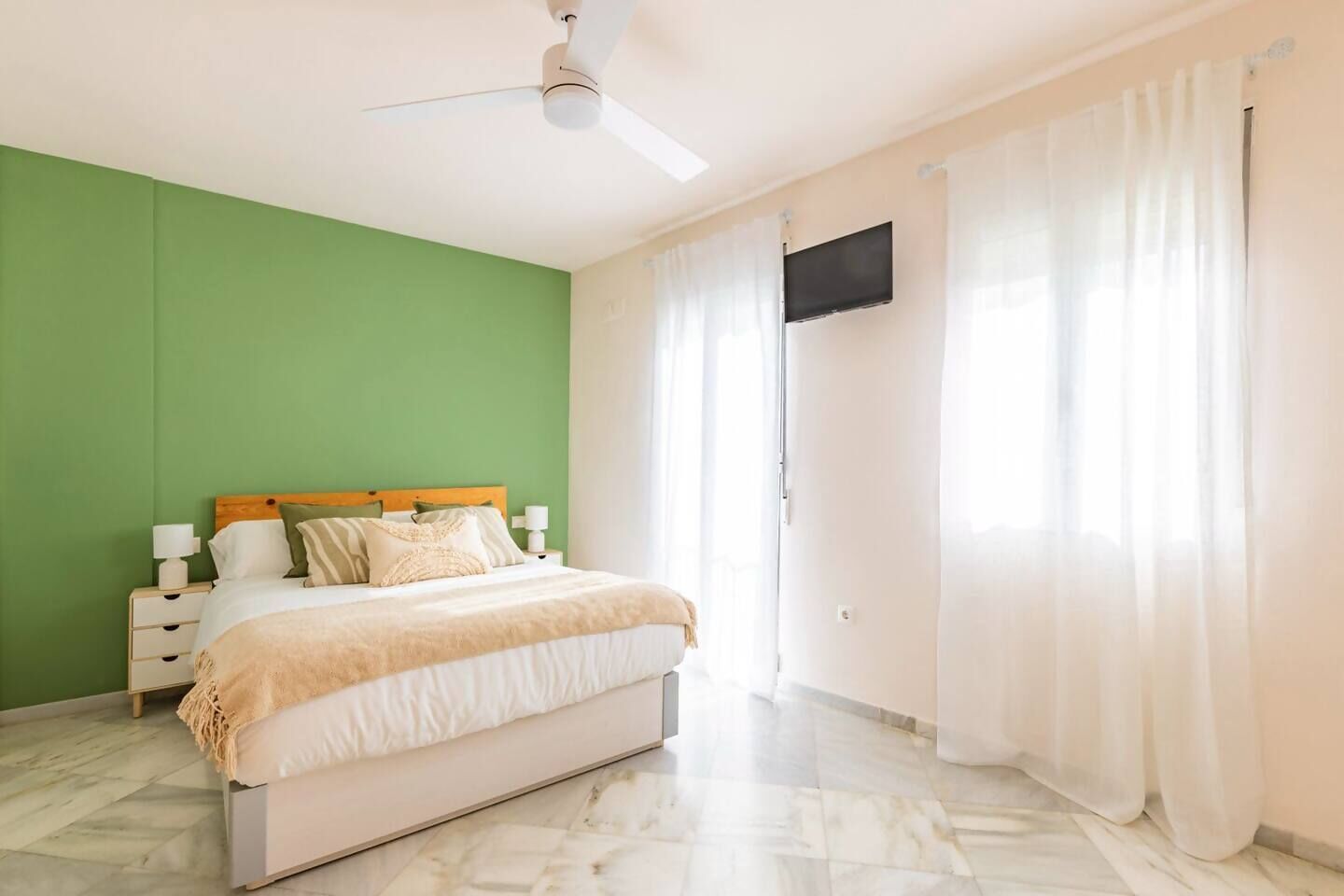 Un dormitorio con cama, ventilador de techo, televisor de pantalla plana y ventana con cortinas.