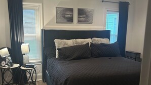 A bedroom with a large bed, a ceiling fan, two wall-mounted picture frames, a bedside table with a lamp, and a window with blinds. - Charming 3-bedroom house in LaPlace with AC, WiFi (LaPlace)