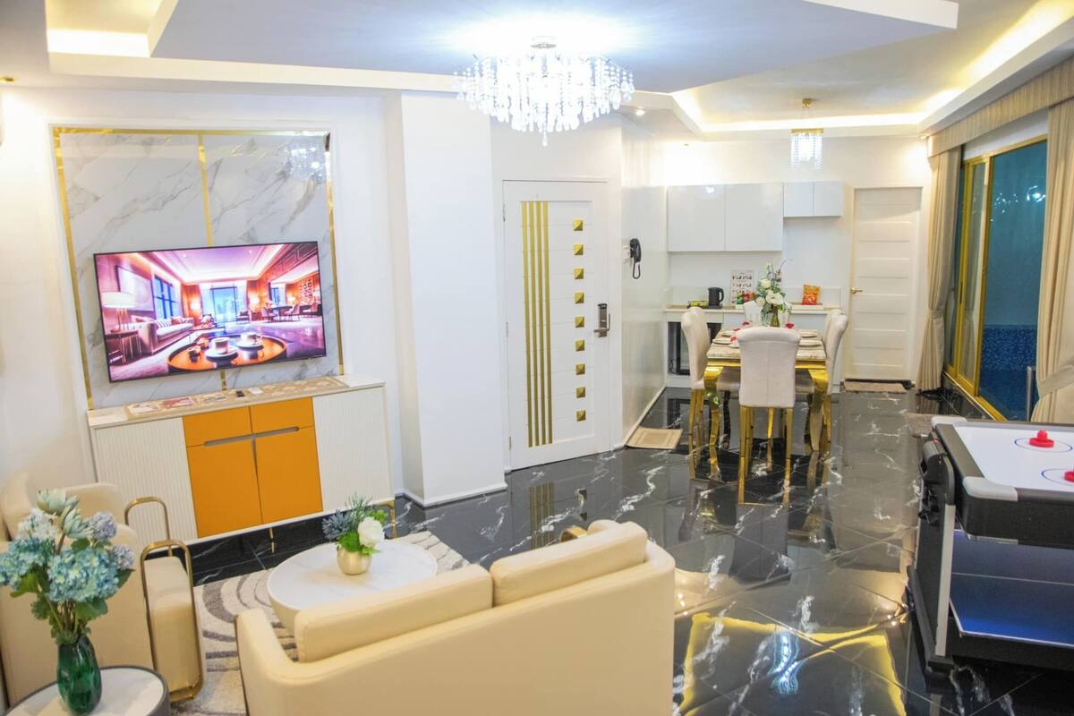 Un salon moderne avec un téléviseur, un canapé et une table basse. Il y a également un coin repas avec une table et des chaises. La pièce est décorée d’un lustre et possède une porte à motifs rayés.
