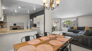 A modern kitchen with white cabinets, a dining area with a wooden table and black chairs, and a gray sofa in the living room. - Desert Dreams Retreat Hot Tub Game Room Fire Pit (Sparks)