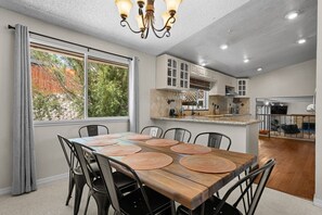 Un espace repas avec une table et des chaises en bois, un lustre et une cuisine en arrière-plan.