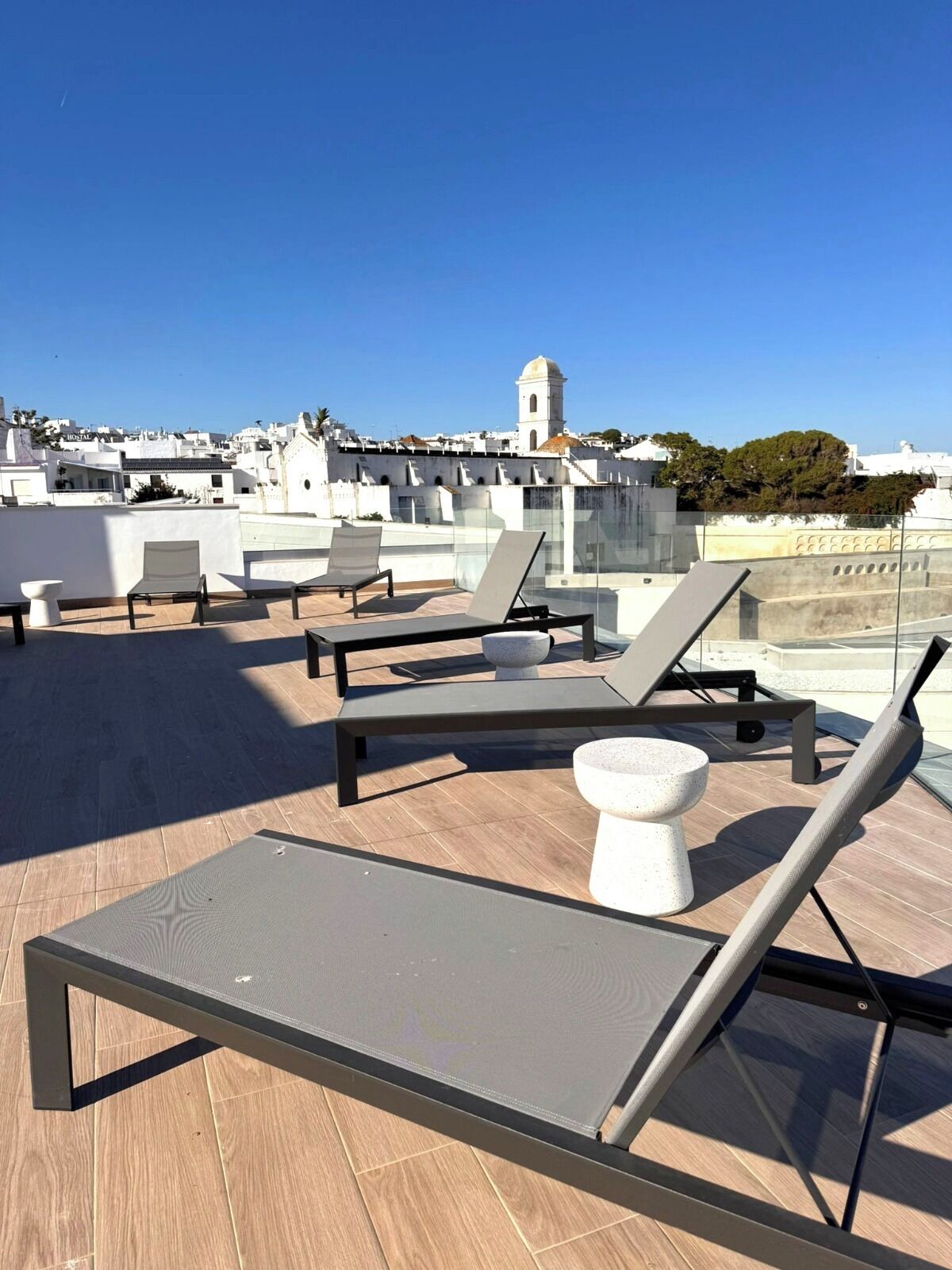 A rooftop terrace with lounge chairs and a stone table, overlooking a cityscape with white buildings and a tower.