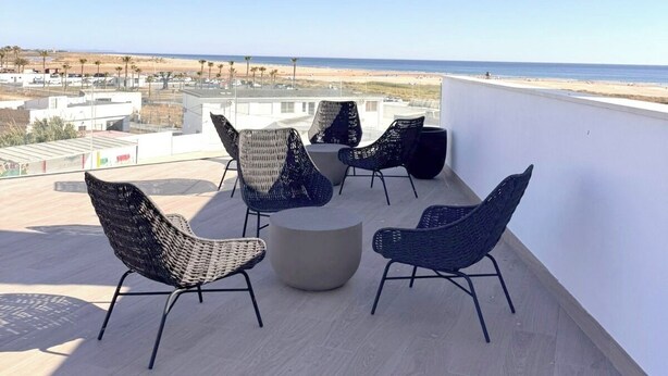 Een terras op het dak met rieten stoelen en een ronde tafel, met uitzicht op een strand en stadsgezicht.