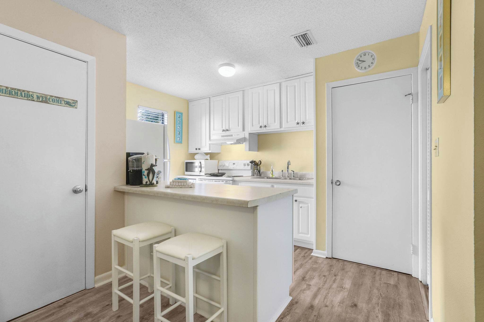 A kitchen with white cabinets, a central island with stools, a microwave, and a clock on the wall.