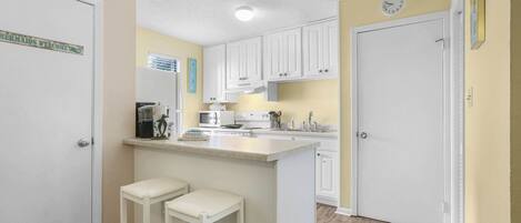 A kitchen with white cabinets, a central island with stools, a microwave, and a clock on the wall.