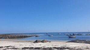 En strand med både i rolige vand, stenet kystlinje og klar, blå himmel.