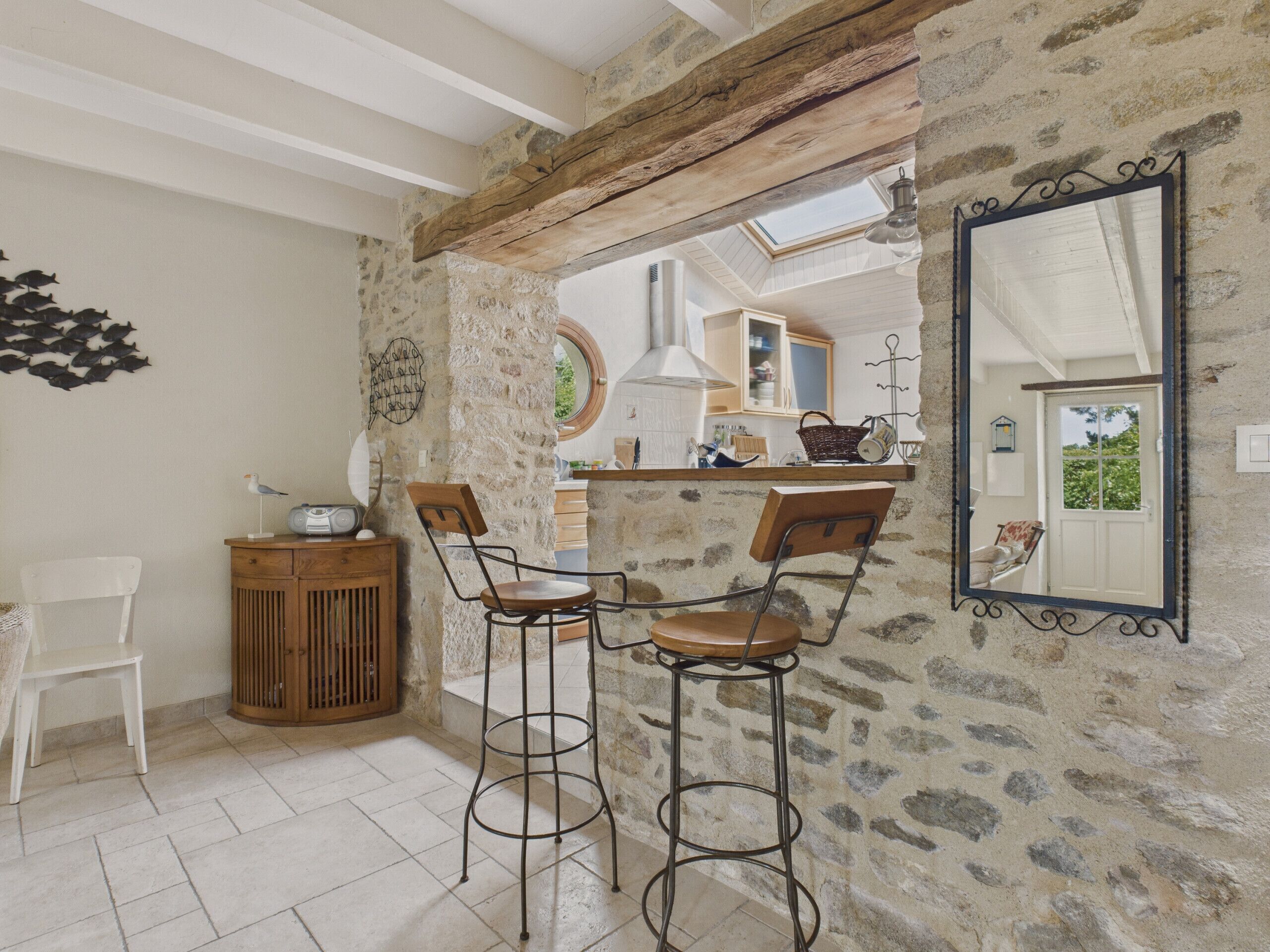 A rustic kitchen with stone walls, wooden beams, and a high ceiling. There are two bar stools, a wooden cabinet, and a white chair. A large mirror hangs on the wall, reflecting the kitchen and a door.