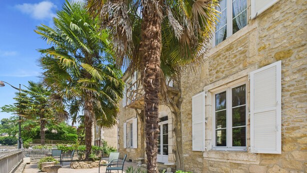 A stone building with white shutters, a palm tree, and outdoor seating.