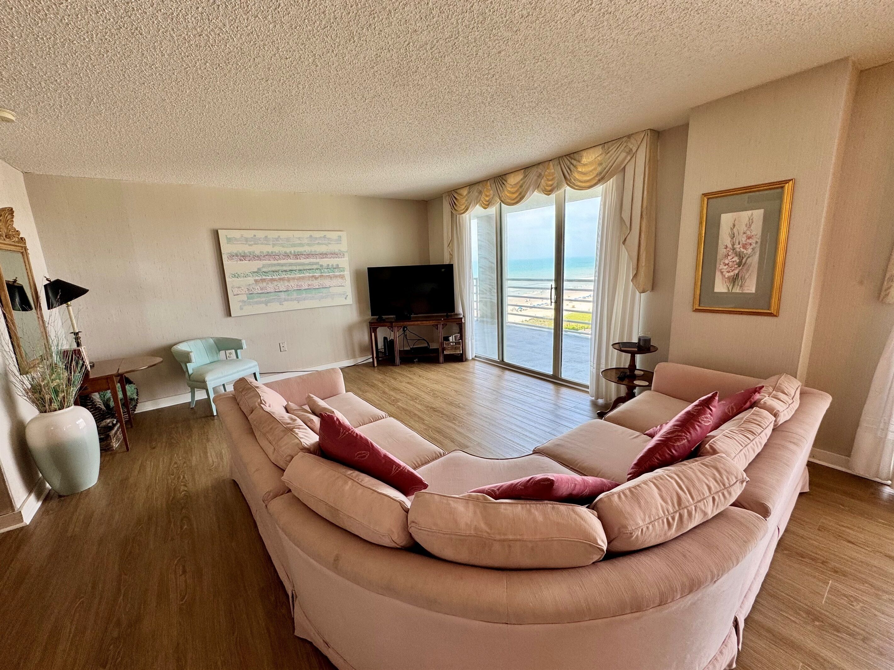 Un salon comprenant un grand canapé d’angle beige, un téléviseur et une grande fenêtre donnant sur la plage.
